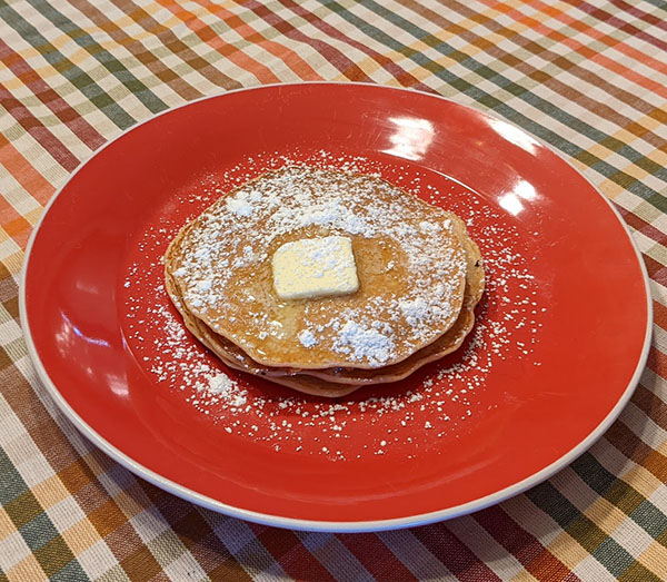 Rusk Pancakes with butter and powdered sugar on a red plate, on top of a plaid tablecloth.